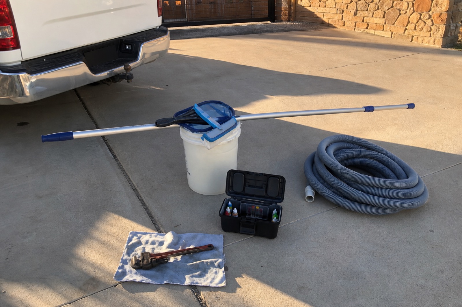 Sunbeam Pools service tools staged on a driveway — telescoping pole on a five-gallon bucket, open test kit case, coiled vacuum hose, pipe wrench on a shop towel, work truck tailgate at the edge of frame.