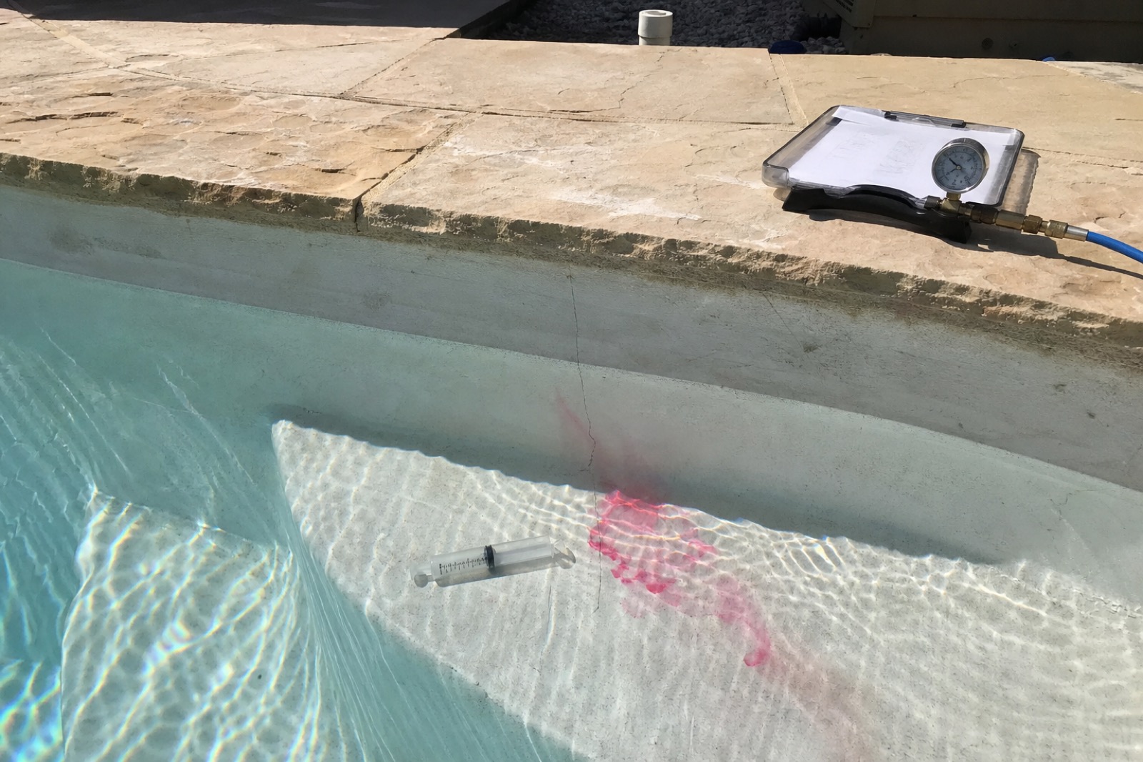 A red dye test trailing into a hairline crack in the plaster of a pool wall, with a pressure-test gauge on the deck above.